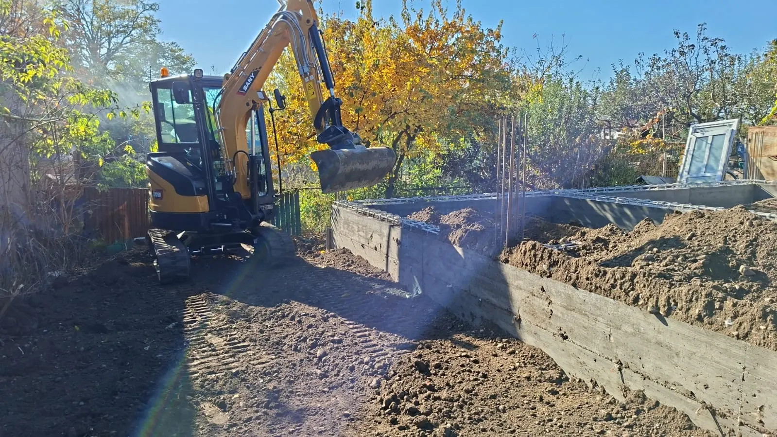 Miniexcavator săpând fundația unei case, cu cofraj de beton turnat și armătură pregătită pe un teren de toamnă.