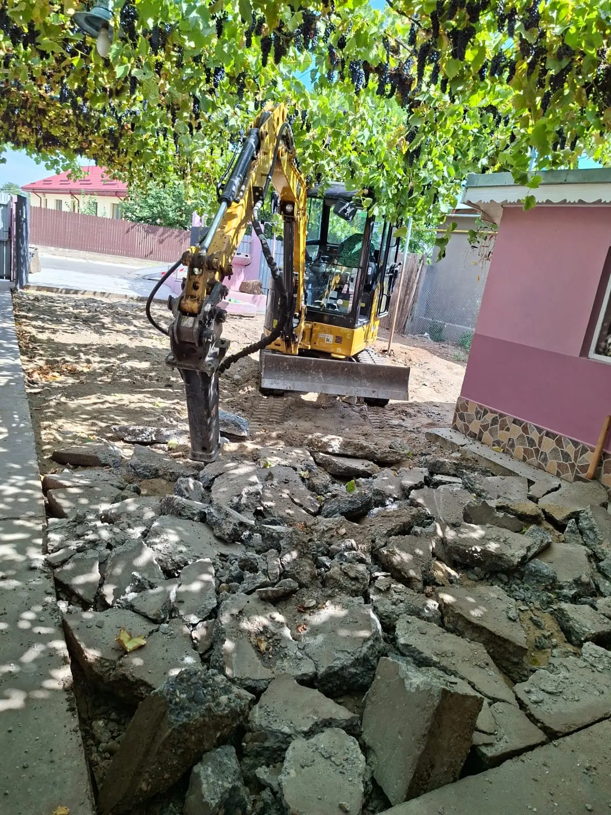 Miniexcavator cu picon hidraulic spărgând o platformă de beton între case, cu molozul adunat pentru evacuare.