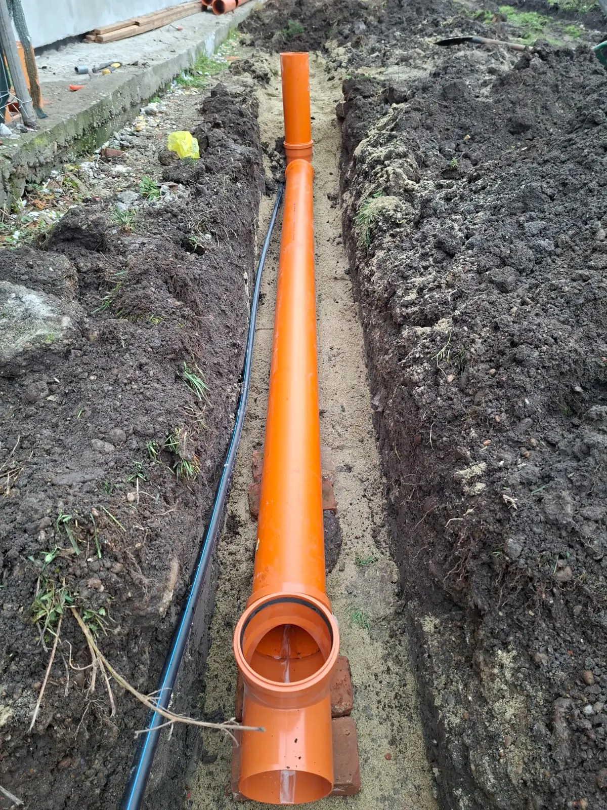 Coloană verticală de canalizare PVC portocalie montată într-o tranșee, pregătită pentru racord.