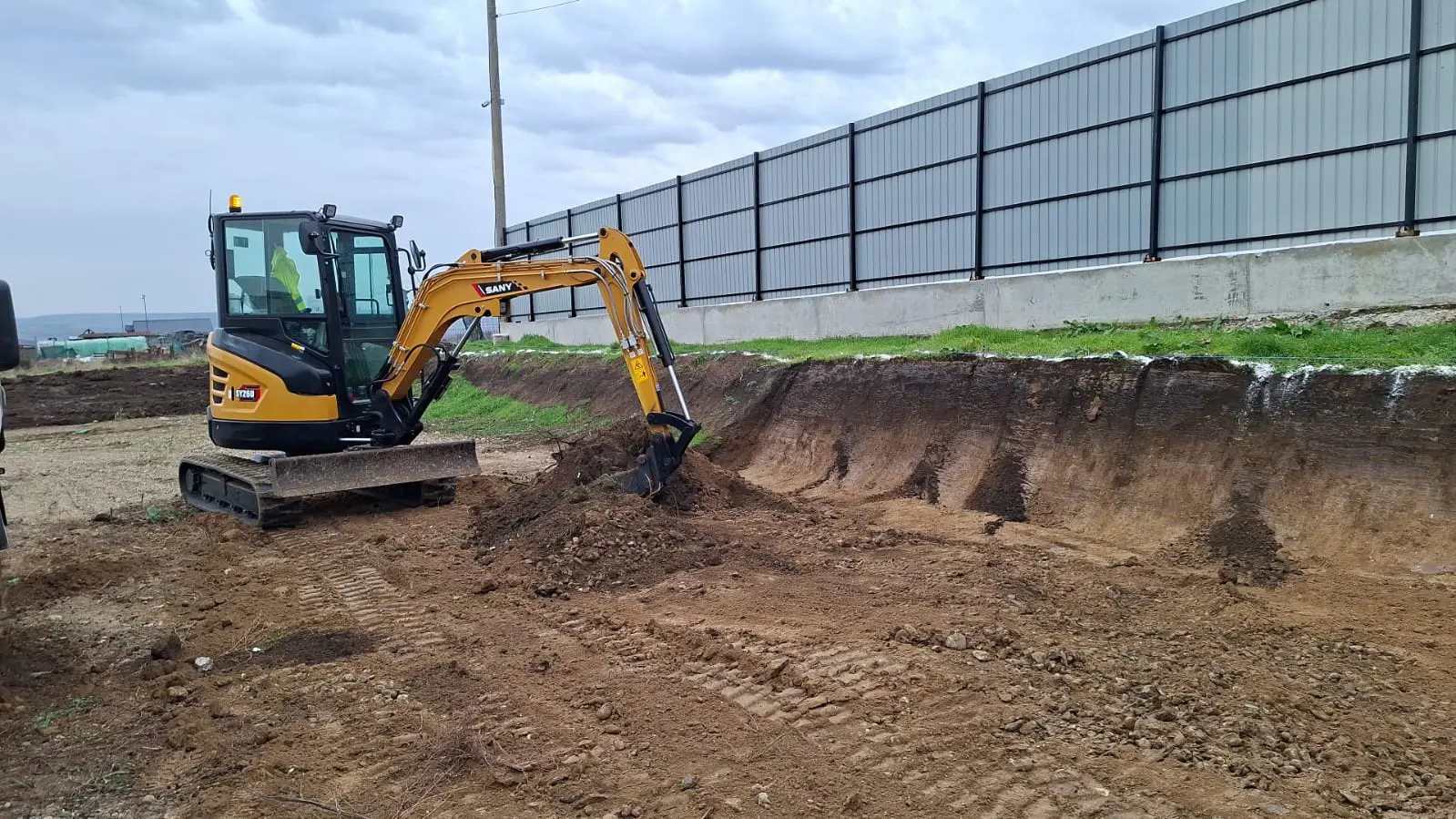 Excavator nivelând un teren lângă un gard de panouri, cu suprafața adusă la cotă.