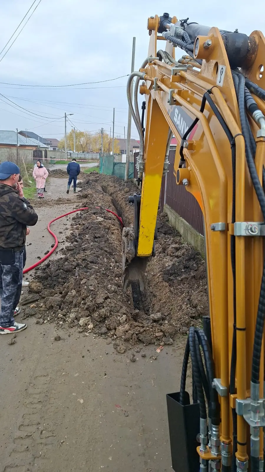 Excavator săpând o tranșee pentru cablu electric pe marginea unei străzi, cu traseul marcat.