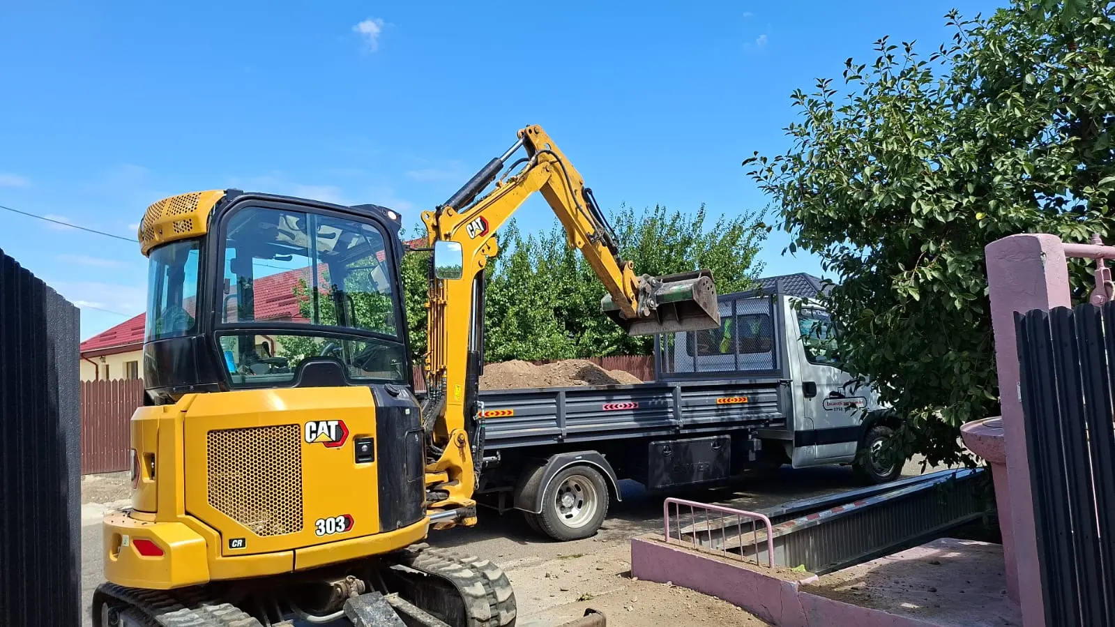 Excavator CAT încărcând molozul într-un autocamion basculant, pe o curte îngustă.
